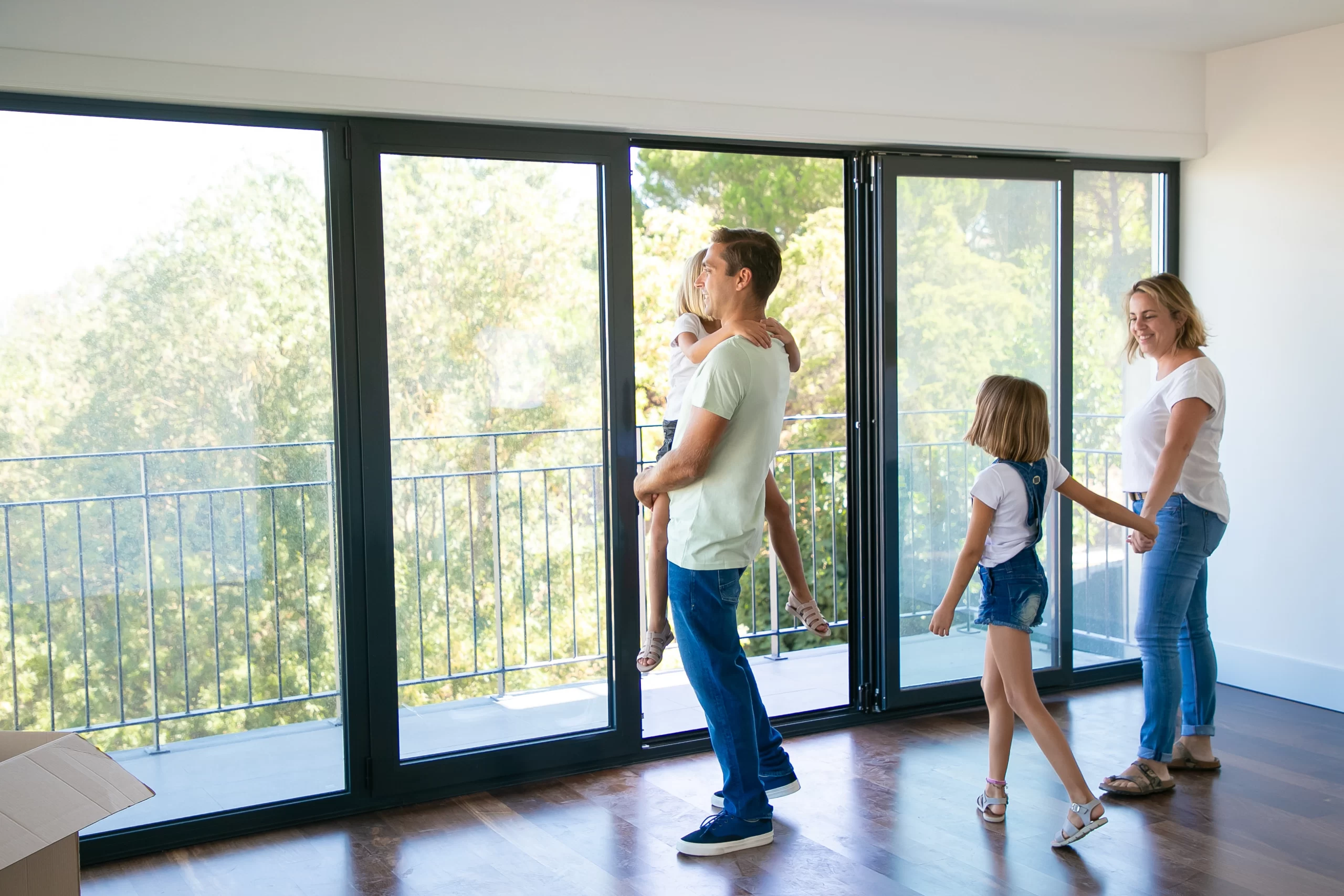 A family stands in a sunlit room in front of expansive black-framed Windows overlooking green trees.