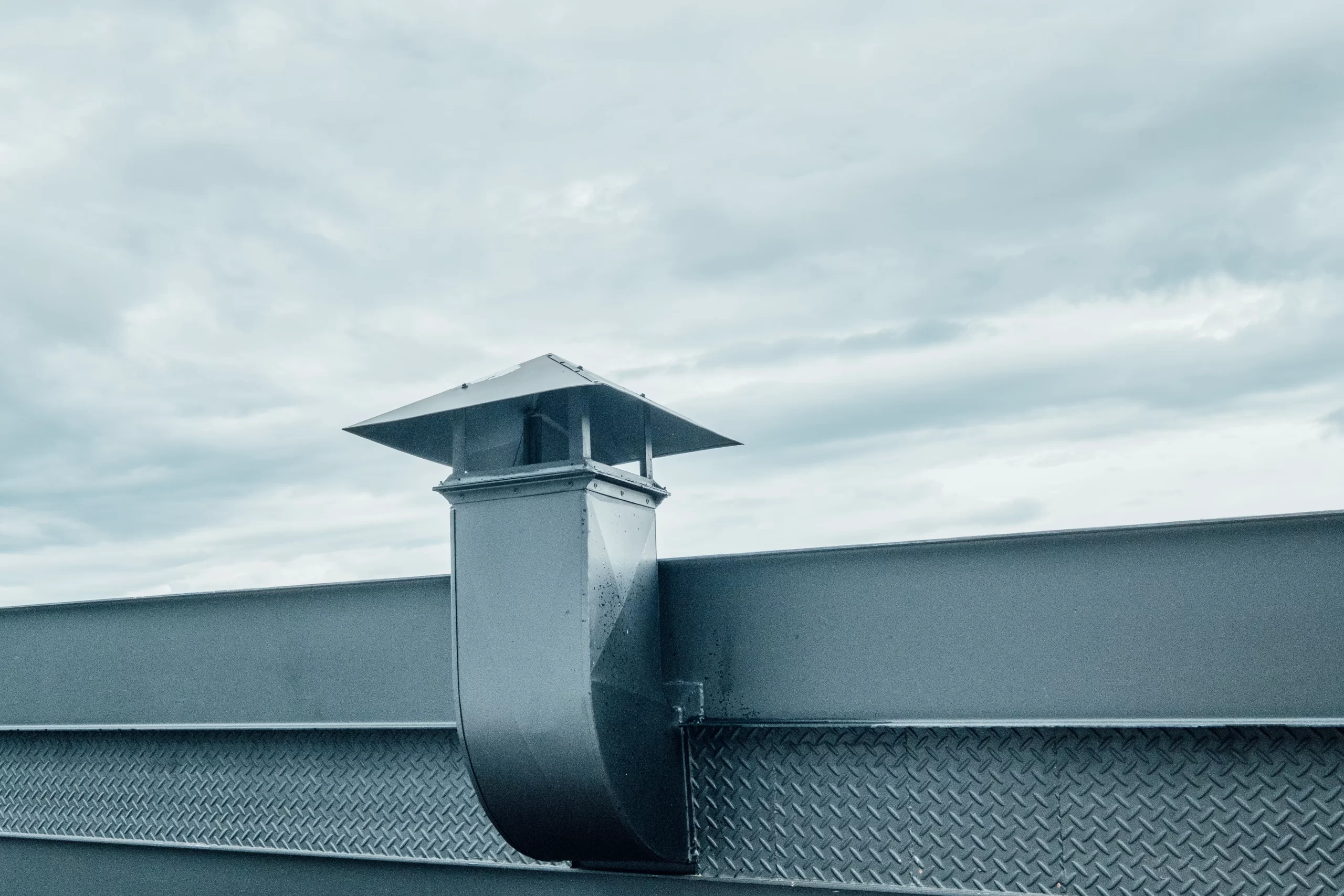 A gray metal vent pipe with a square cap stands against a cloudy sky next to a Gutter system.