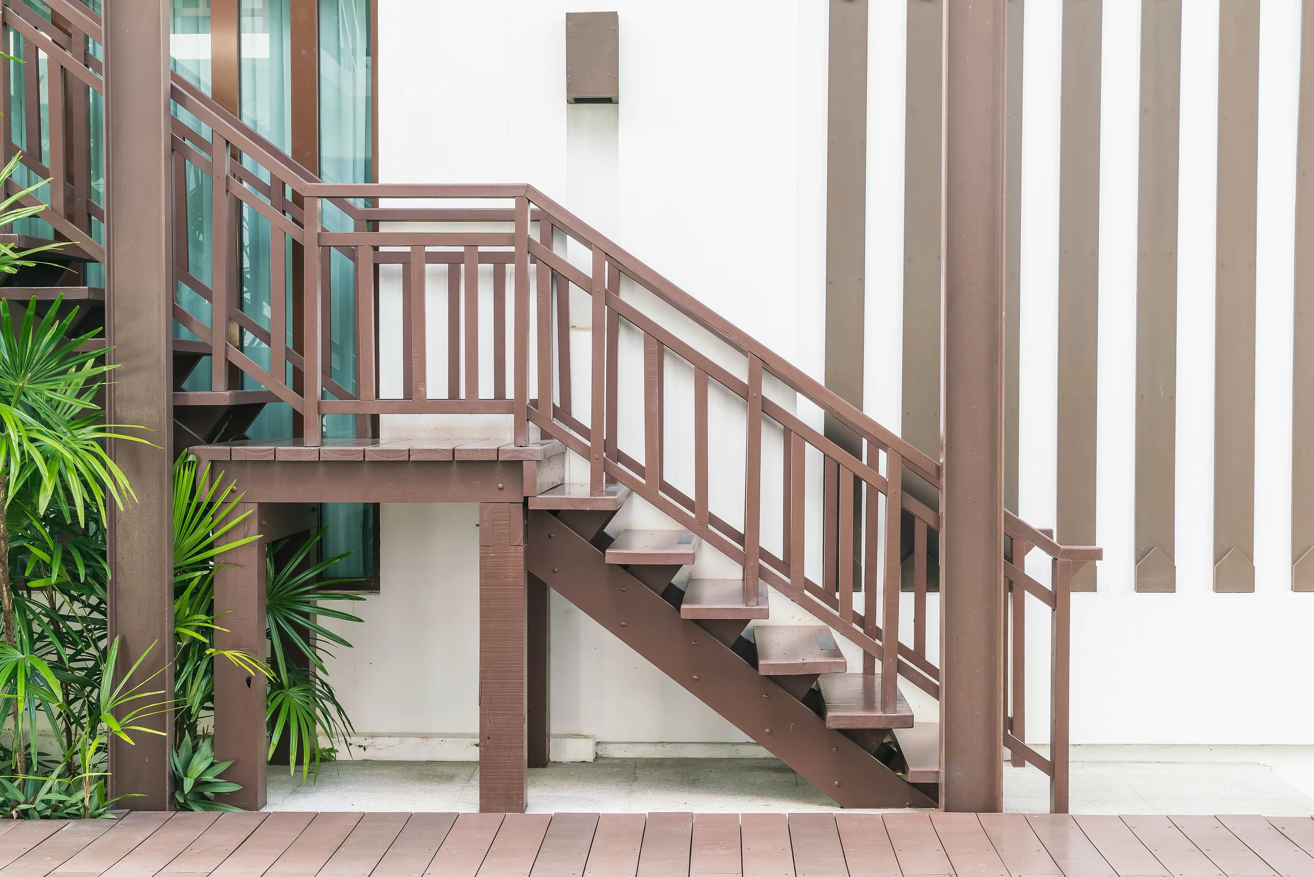 A brown wooden staircase with brown Metal Rails leads to a balcony against a white slatted wall.