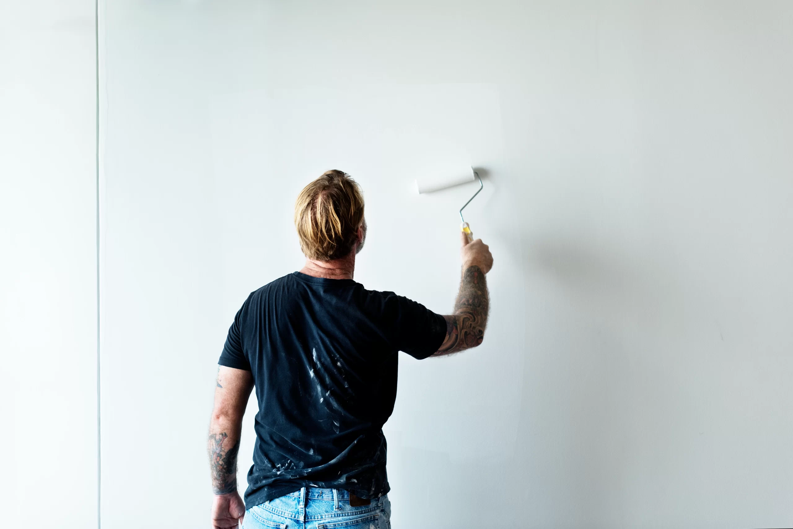 A tattooed man in a black t-shirt uses a roller to apply fresh white Paint to a large indoor wall.