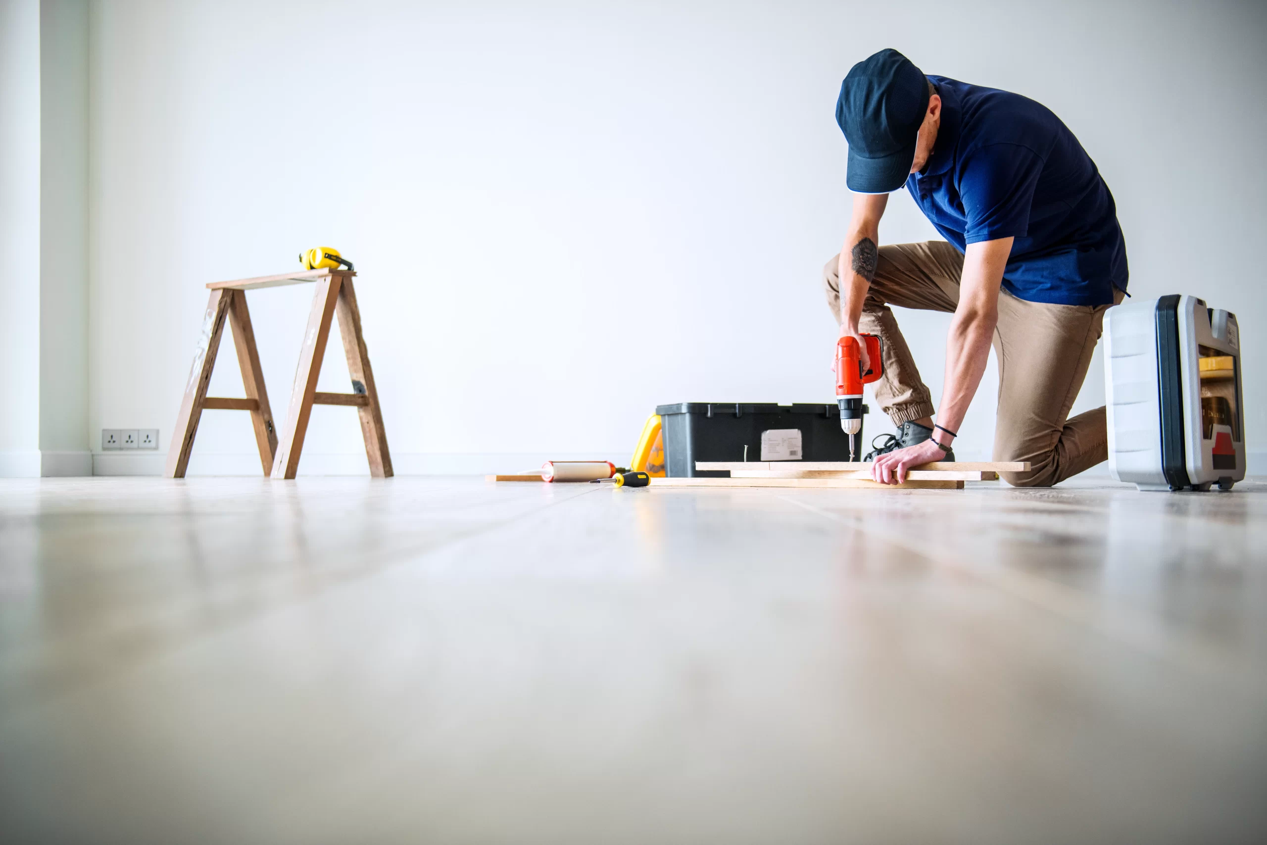 A worker in a blue polo shirt kneels on a bright room floor to drill into a light-colored flooring plank.