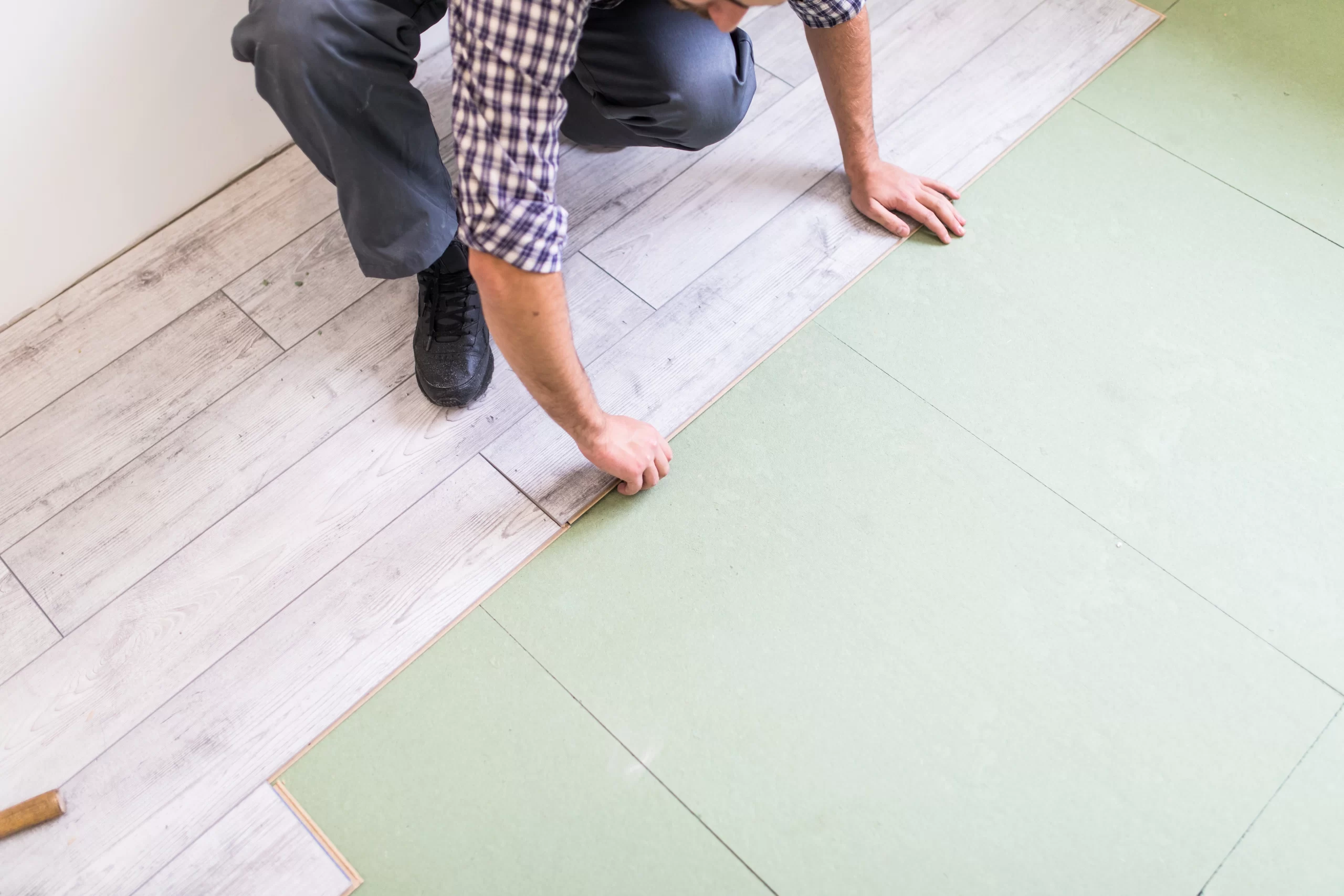 A worker in a plaid shirt carefully aligns a grey wood-grain flooring plank over a green underlayment.