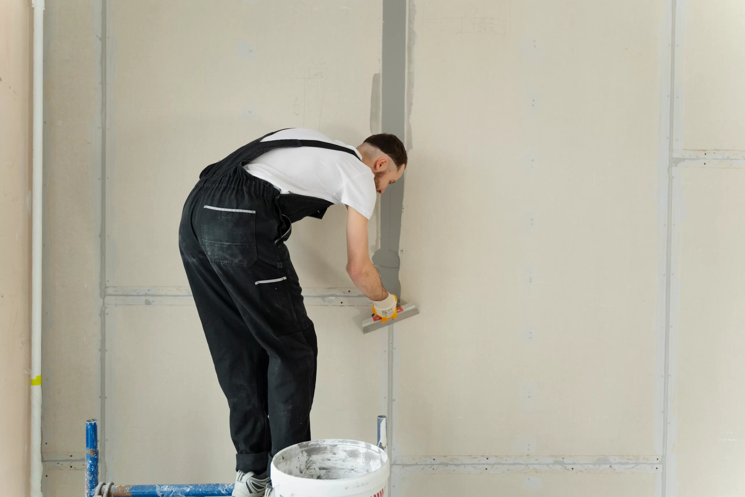 A worker in black overalls uses a trowel to apply joint compound to a Drywall seam in a new room.