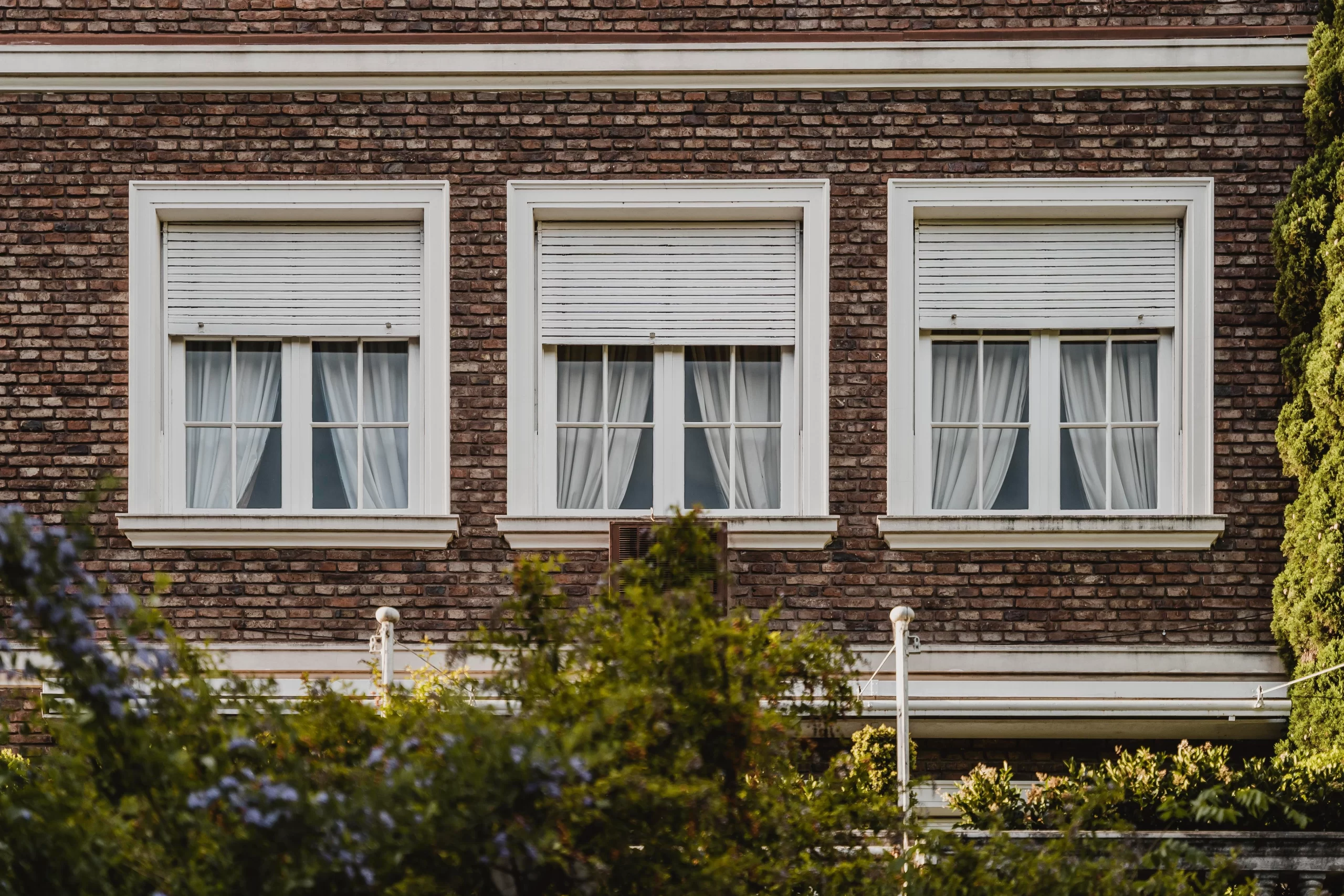 Three white sash Windows with roller shutters set into a brown brick wall above green garden bushes.