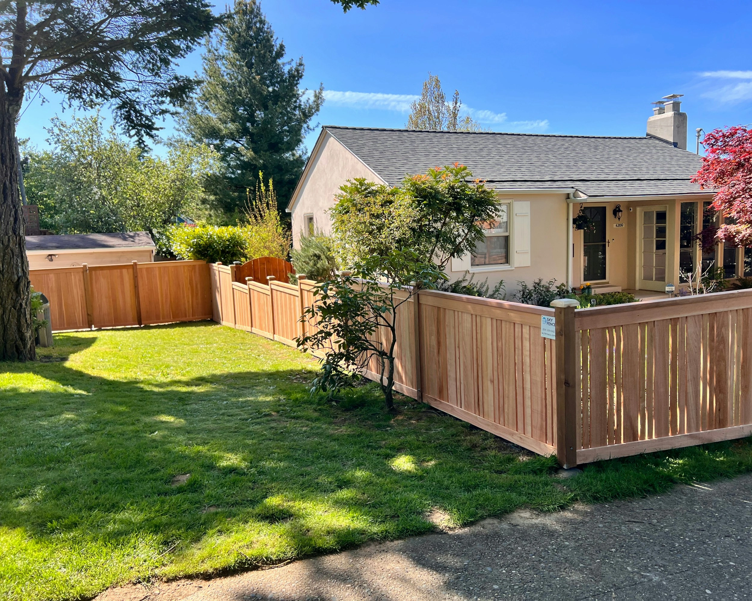 A neat wooden Fencing enclosure surrounds a lush green lawn and a small beige house under a clear blue sky.