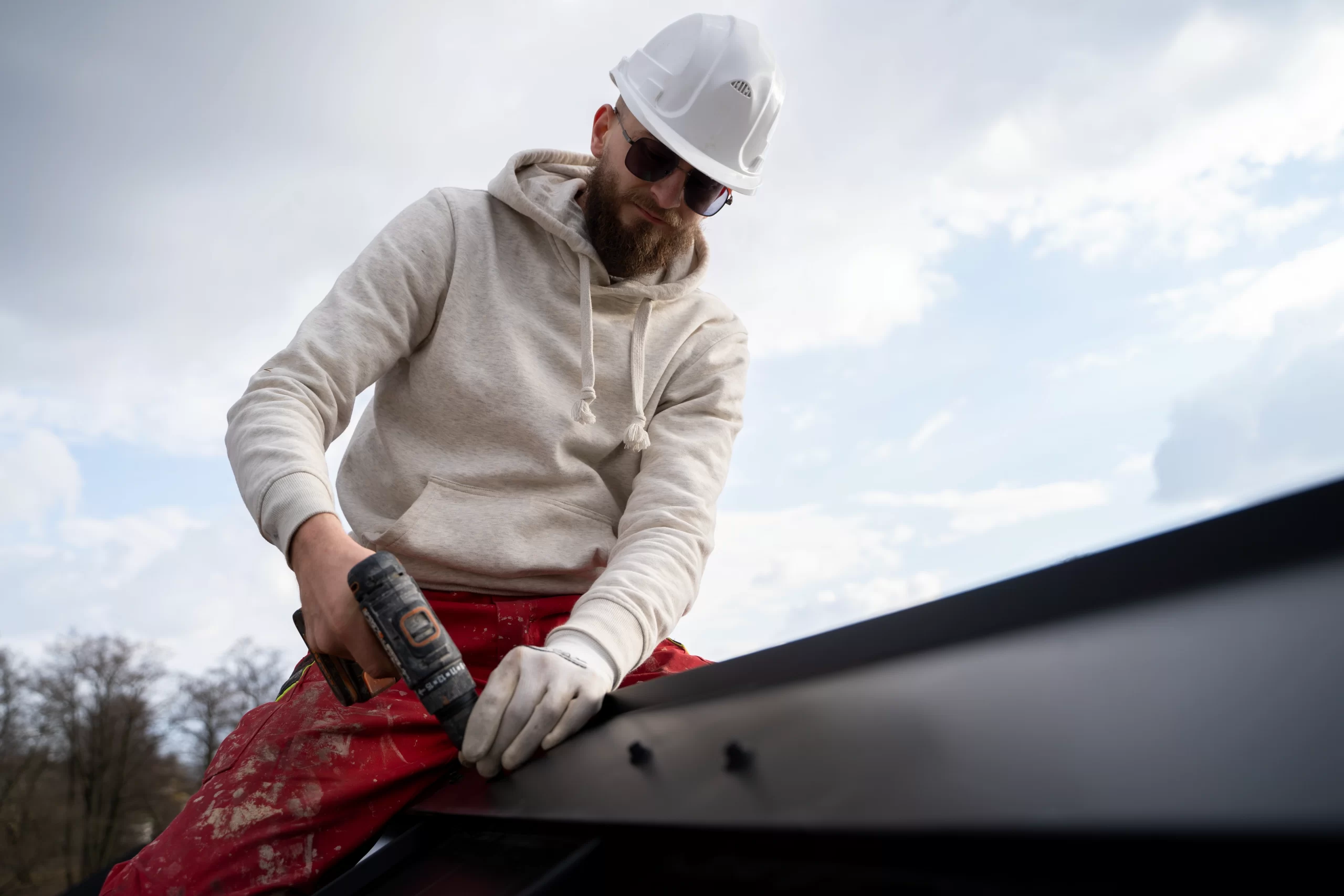 A worker in a white hard hat and sunglasses uses a drill to secure metal roofing near a black Gutter.