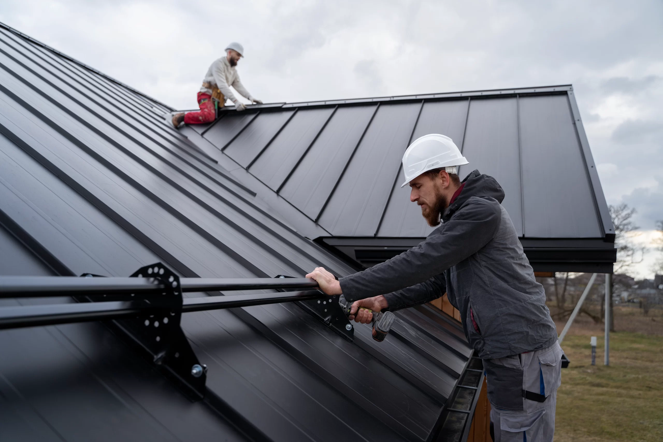 Roofer in a white hard hat securing a black metal rail to the standing seam of a roofing structure with a drill