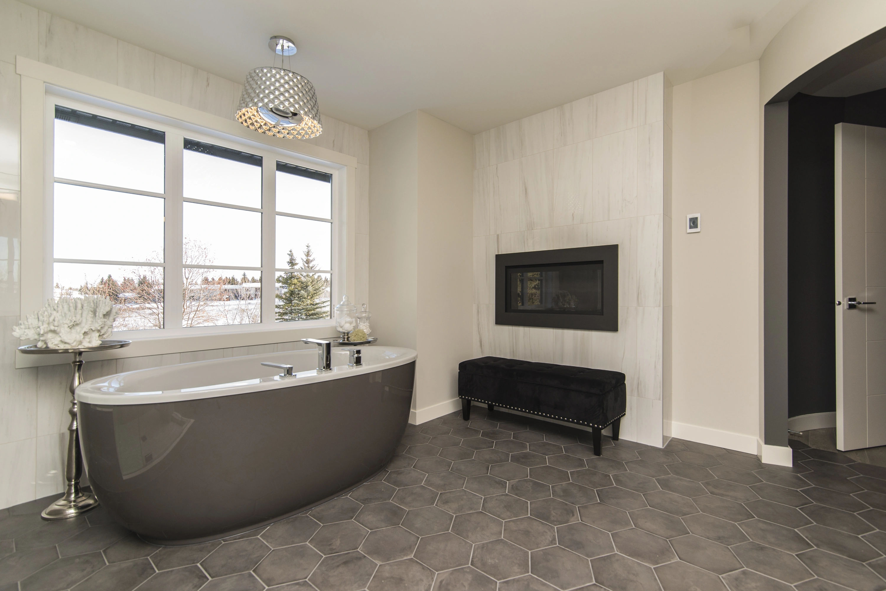 A freestanding grey bathtub sits on dark grey hexagonal floor Tile near a fireplace and large window.
