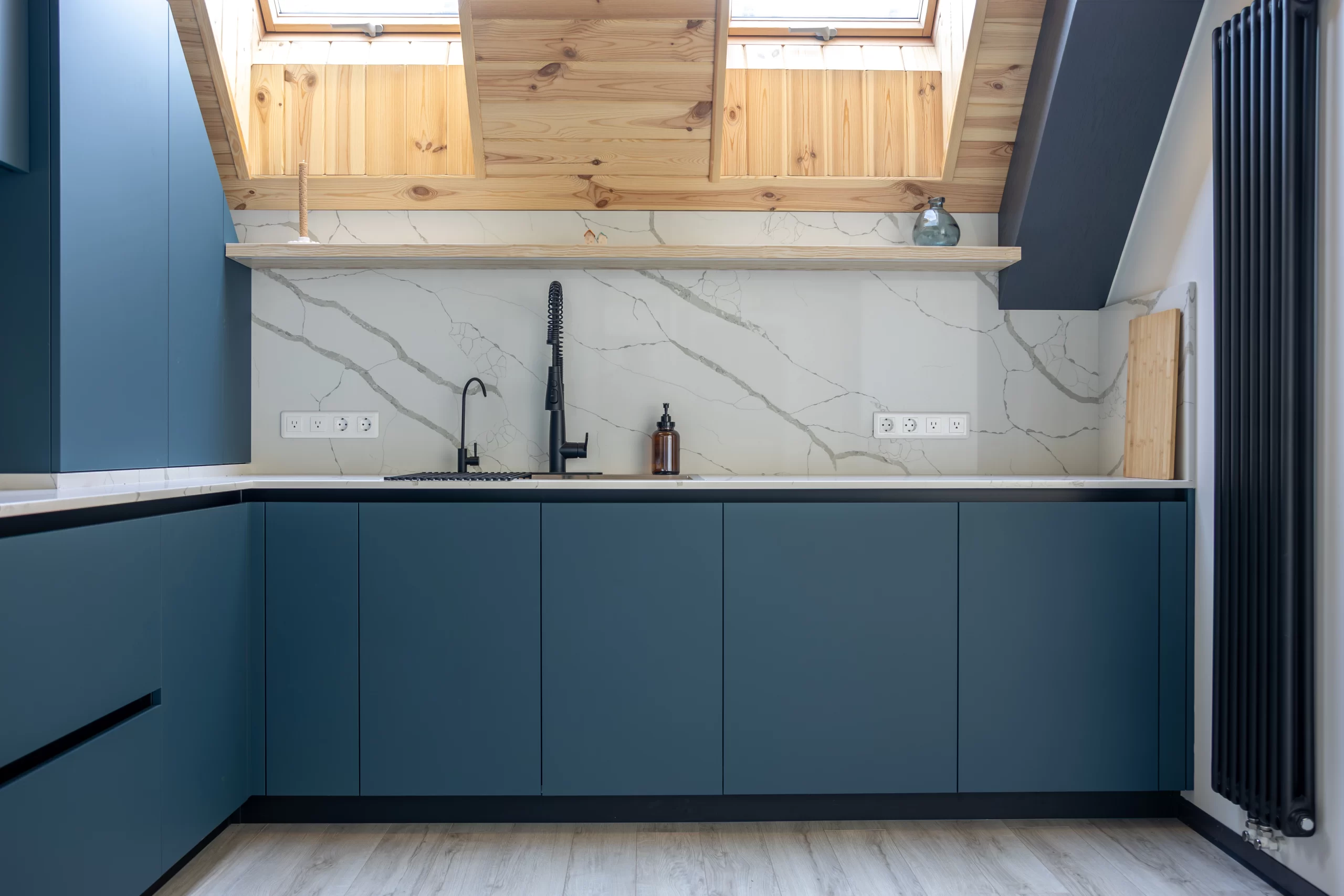 Matte blue handleless Cabinets under a marble backsplash and slanted wooden ceiling with skylights.
