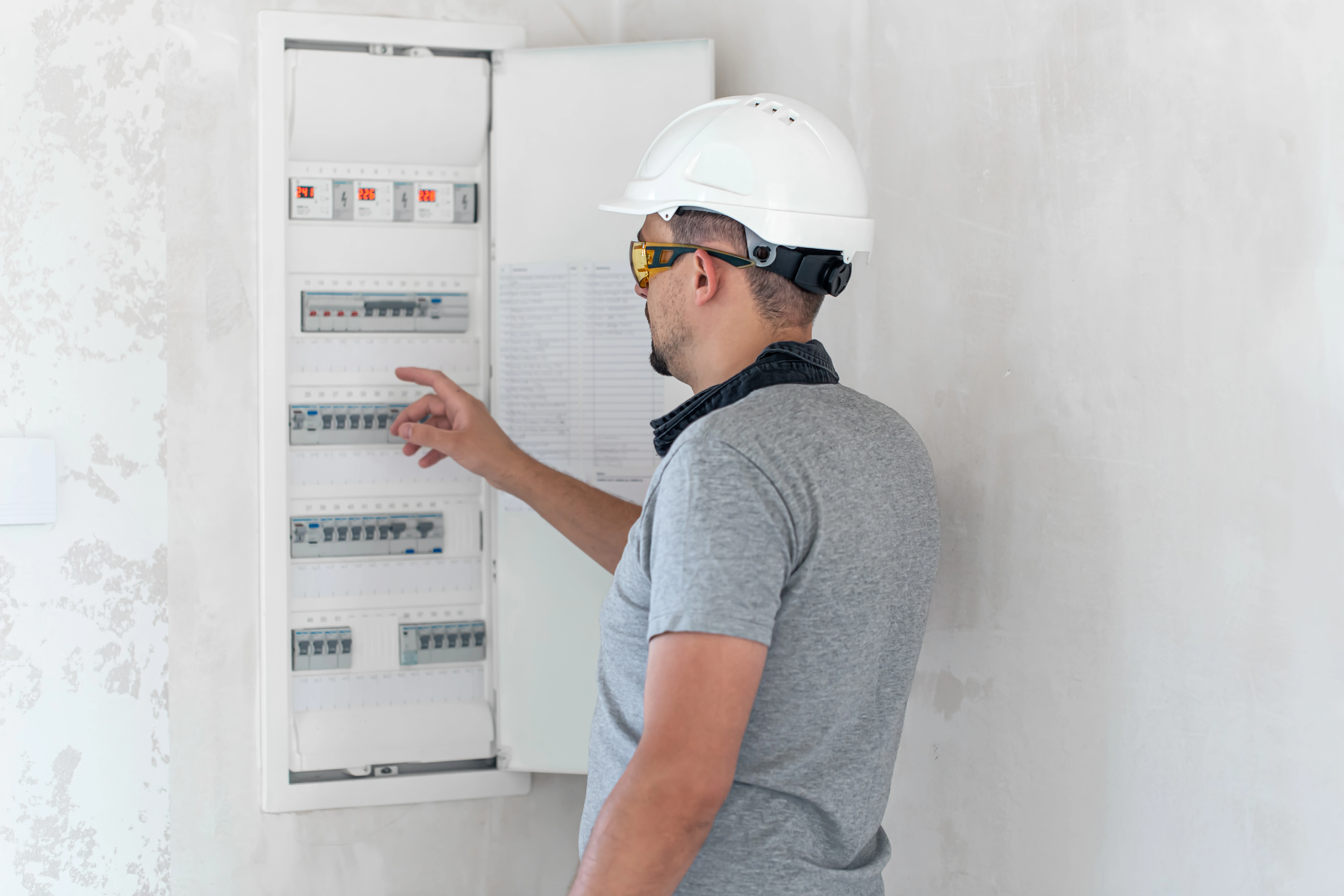 A worker in a hard hat examines the circuit breakers in a white wall-mounted Electrical panel.