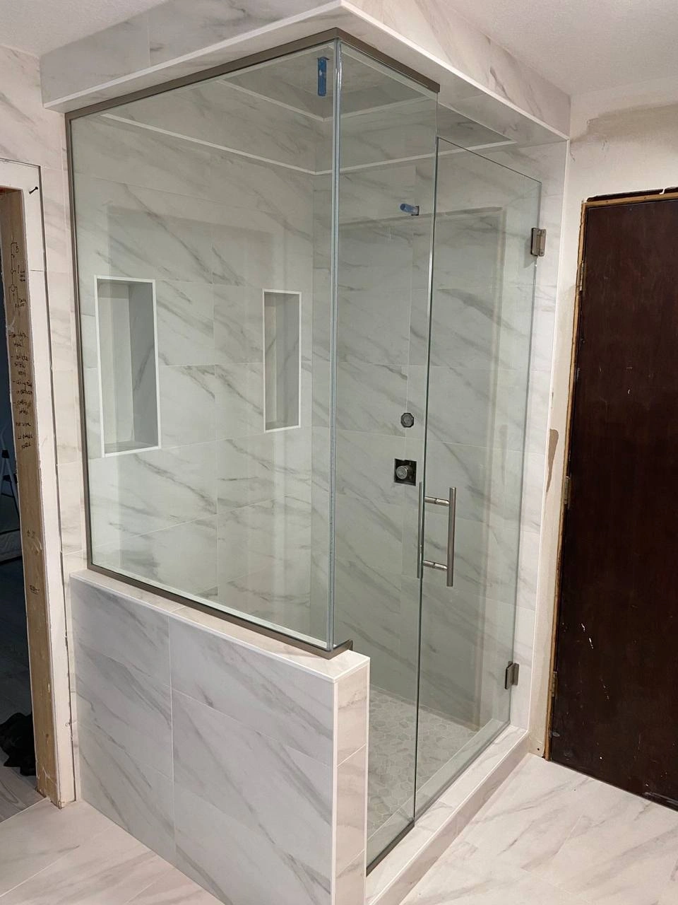 A corner shower unit with large marble tiles and clear Shower Glass Doors installed on a pony wall.