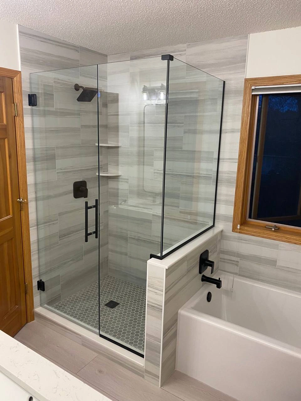 Corner walk-in shower with matte black trim and Shower Glass Doors next to a white soaking tub.